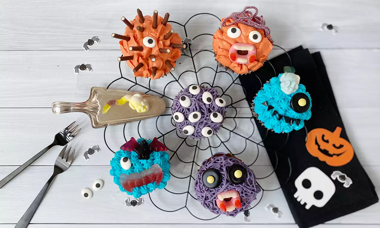 Bunte Halloween-Cupcakes in Monstergestalt mit Augen, Tentakeln und Dekoration auf einem schwarzen Kuchengitter und stilvollem Besteck drapiert