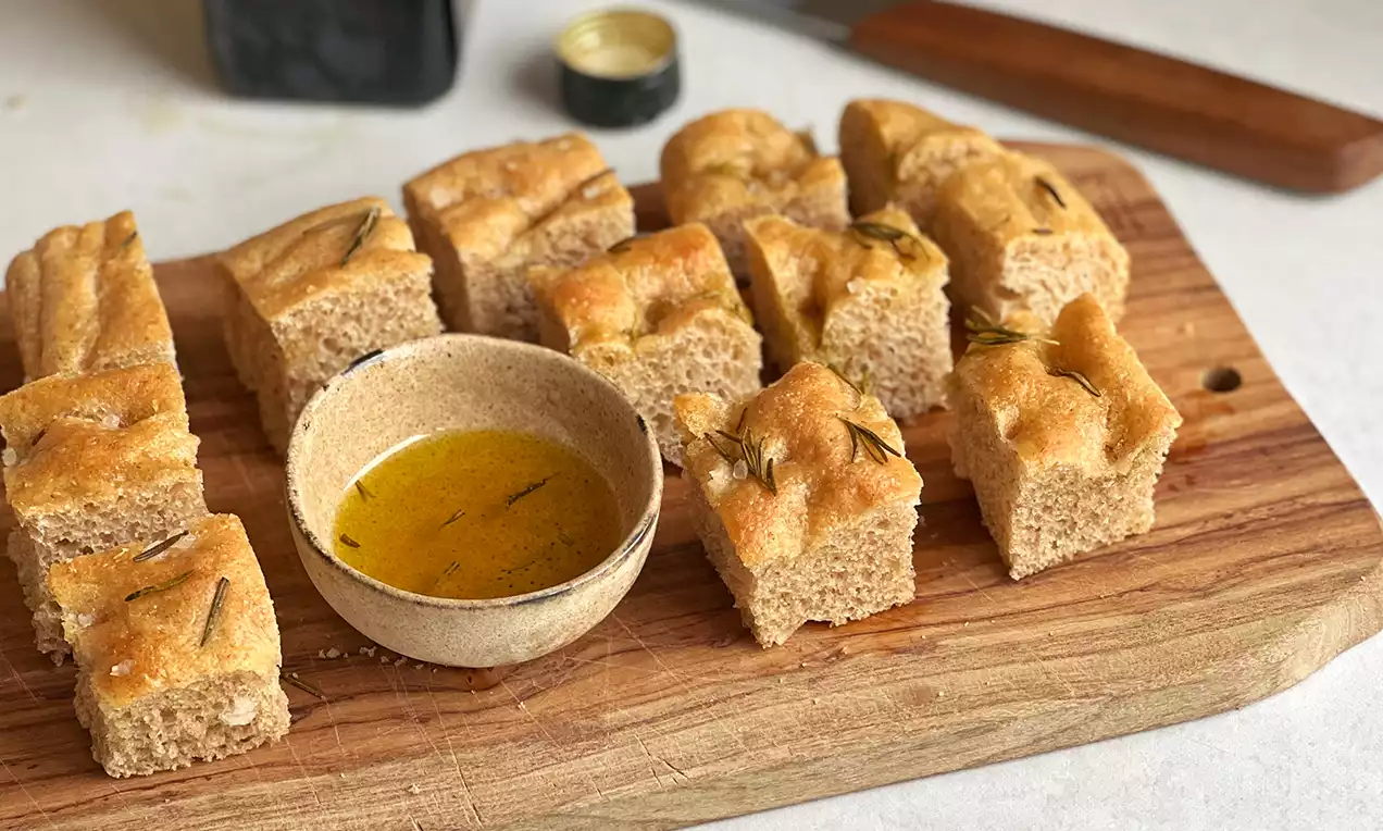 Rosmarin Focaccia mit goldbrauner Kruste in rechteckige Stücke geschnitten, mit knuspriger Textur neben einer Schale mit Olivenöl auf Holzbrett