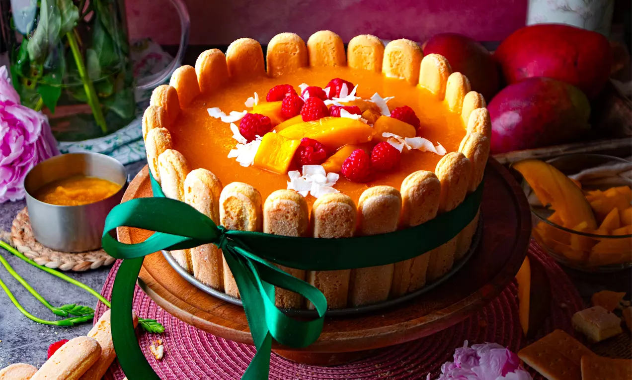 Бисквитена торта с яркооранжев крем отгоре, декорирана с резени манго, малини и кокосови стърготини, обгърната с бишкоти и зелената панделка