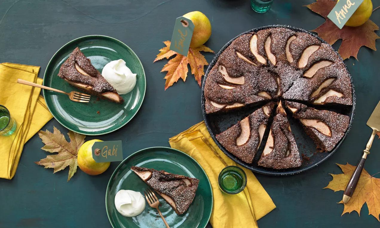 Тъмнокафяв сладкиш с матова текстура, гарниран с сочни резени круши, подреден симетрично върху тъмна чиния, украсен с есенни листа