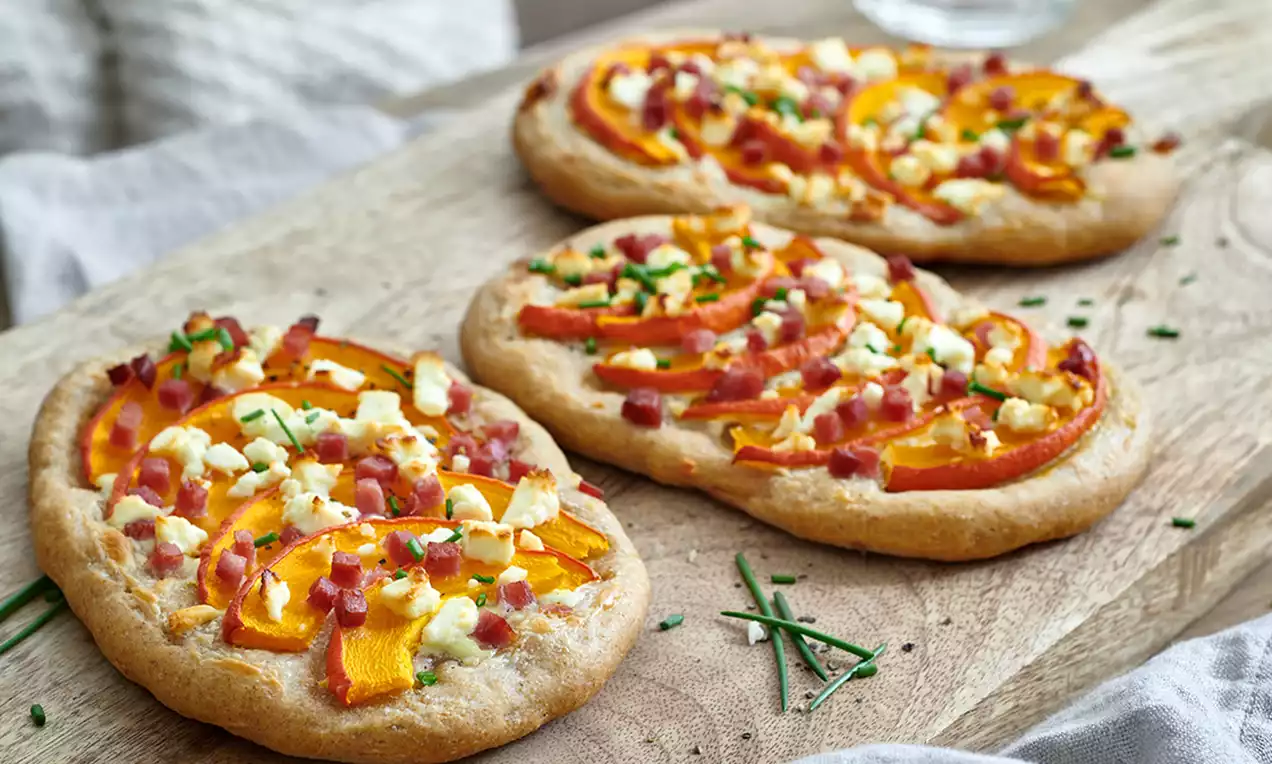 Goldbraune, flache Rahmfladen mit Kürbisstreifen, Speckwürfeln und Krümeln von weißem Käse auf einem rustikalen Holzbrett angerichtet