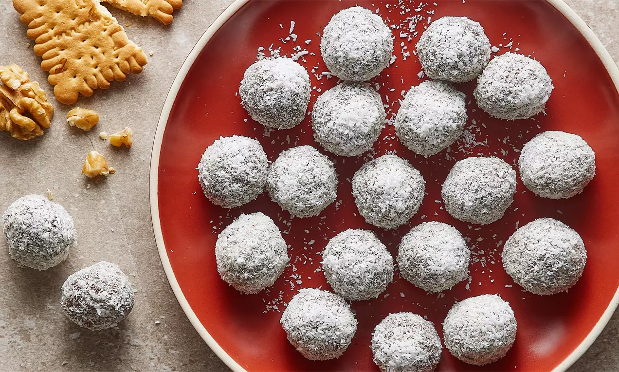 Kırmızı bir tabakta düzenli şekilde sıralanmış, beyaz hindistancevizi kaplı yuvarlak keçiboynuzlu bisküvi topları, yanında ceviz ve bisküvi parçaları