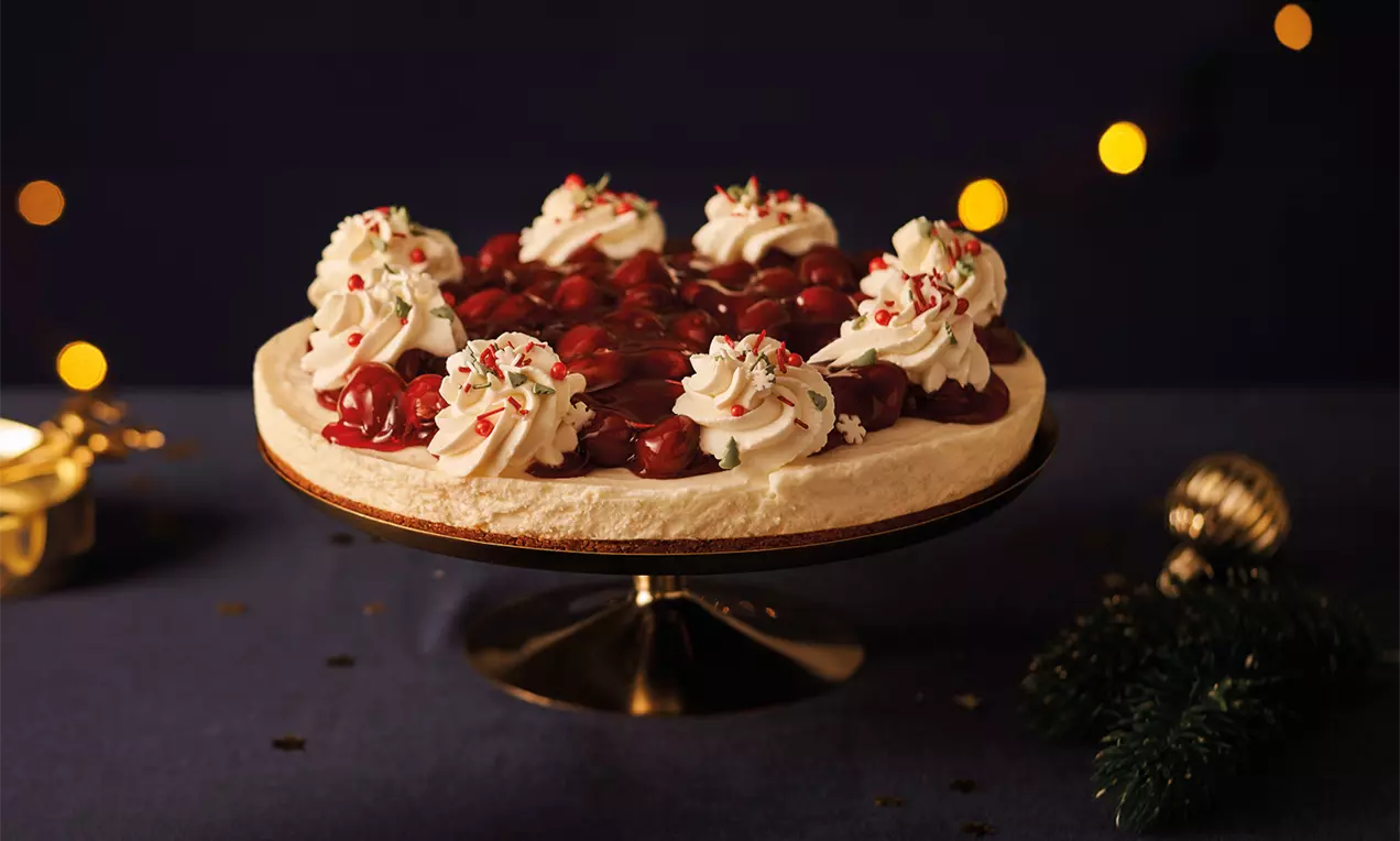 Ronde MonChoutaart met een romige witte vulling, rode glanzende kersen en toefjes slagroom, versierd met sprinkles op een goudkleurige taartstandaard