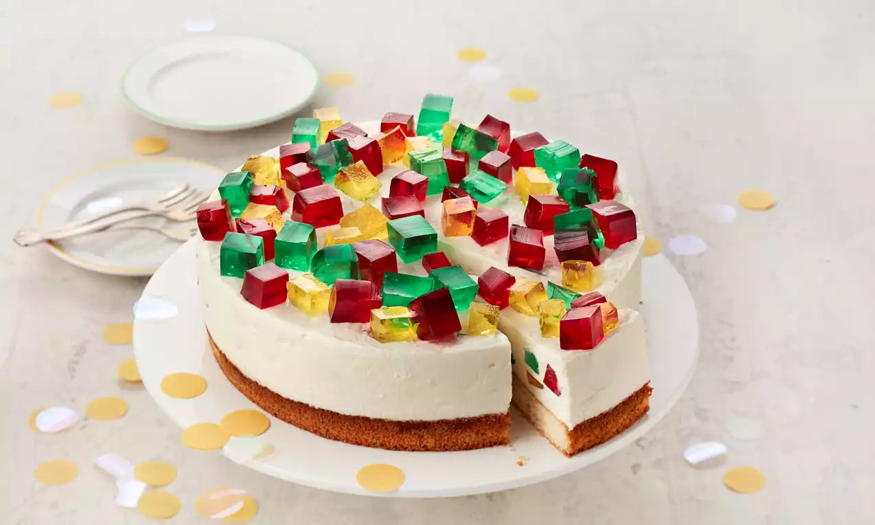 Bunte Würfel aus rotem, grünem und gelbem Wackelpudding als Dekoration auf einer hellen Cremetorte mit braunem Boden auf weißer Platte