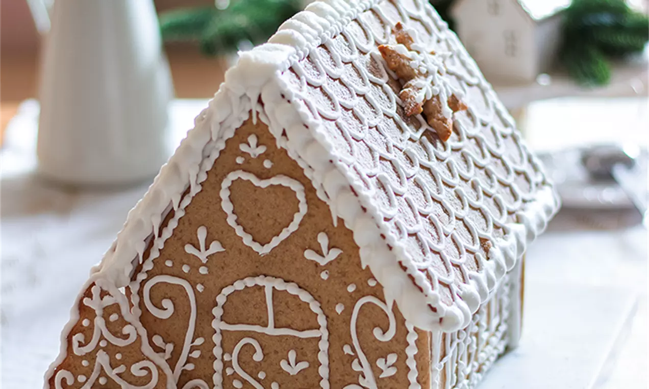 Джинджифилова къщичка с топъл кафяв цвят, декорирана с бял глазур в детайлни орнаменти, на бяла повърхност с фонови зелени клонки