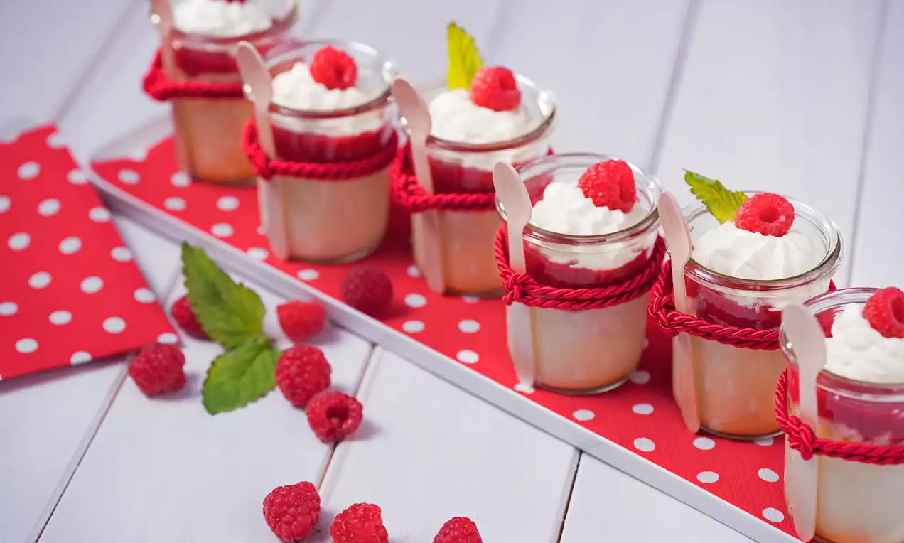 Skutina krema bledo rumene barve v steklenem kozarcu, prekrita z mehko belo smetano in svežimi rdečimi malinami, okrašena z zelenimi lističi na rdeče-beli podlagi