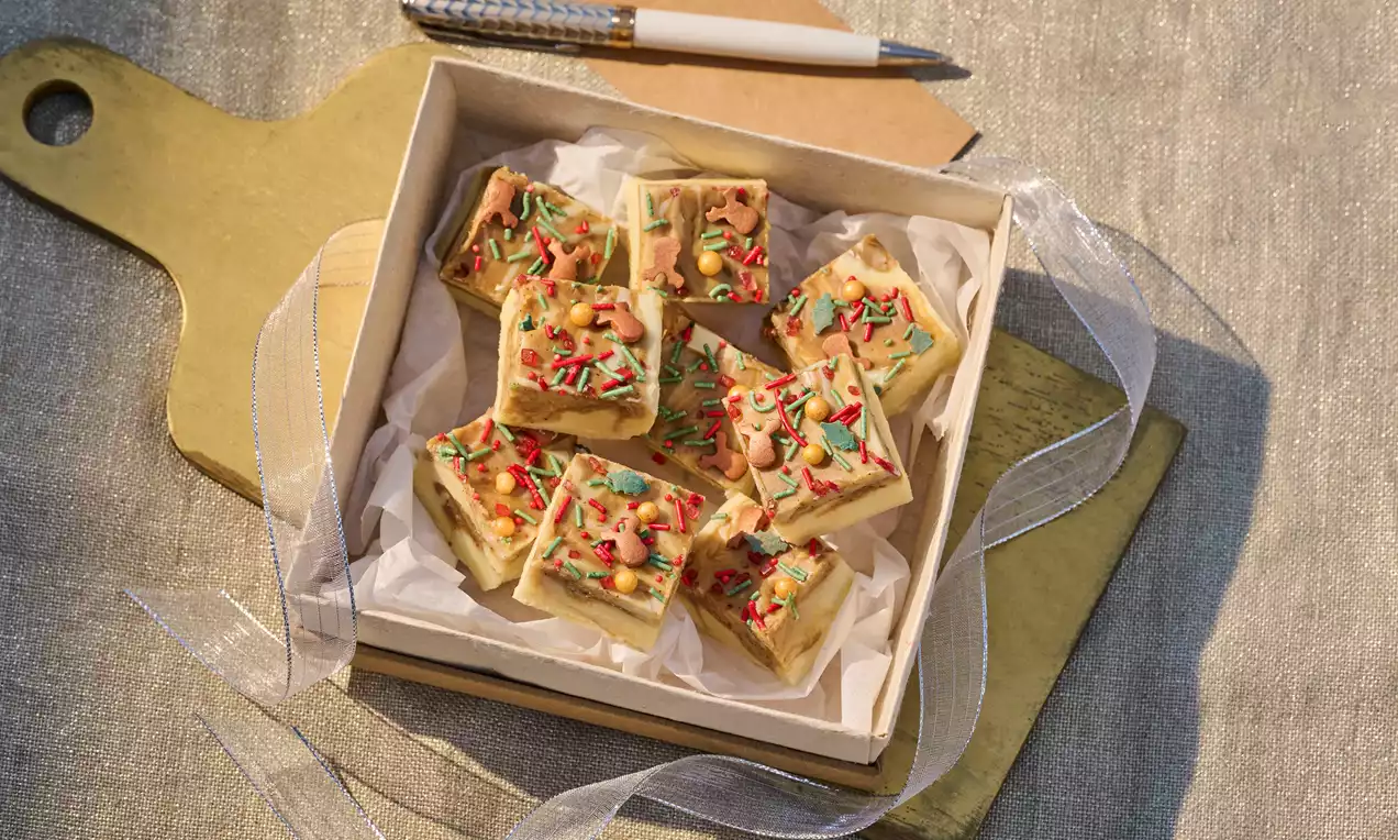 Golden brown fudge squares with a smooth surface topped with green and red sprinkles, arranged neatly in a paper-lined box on a wooden tray