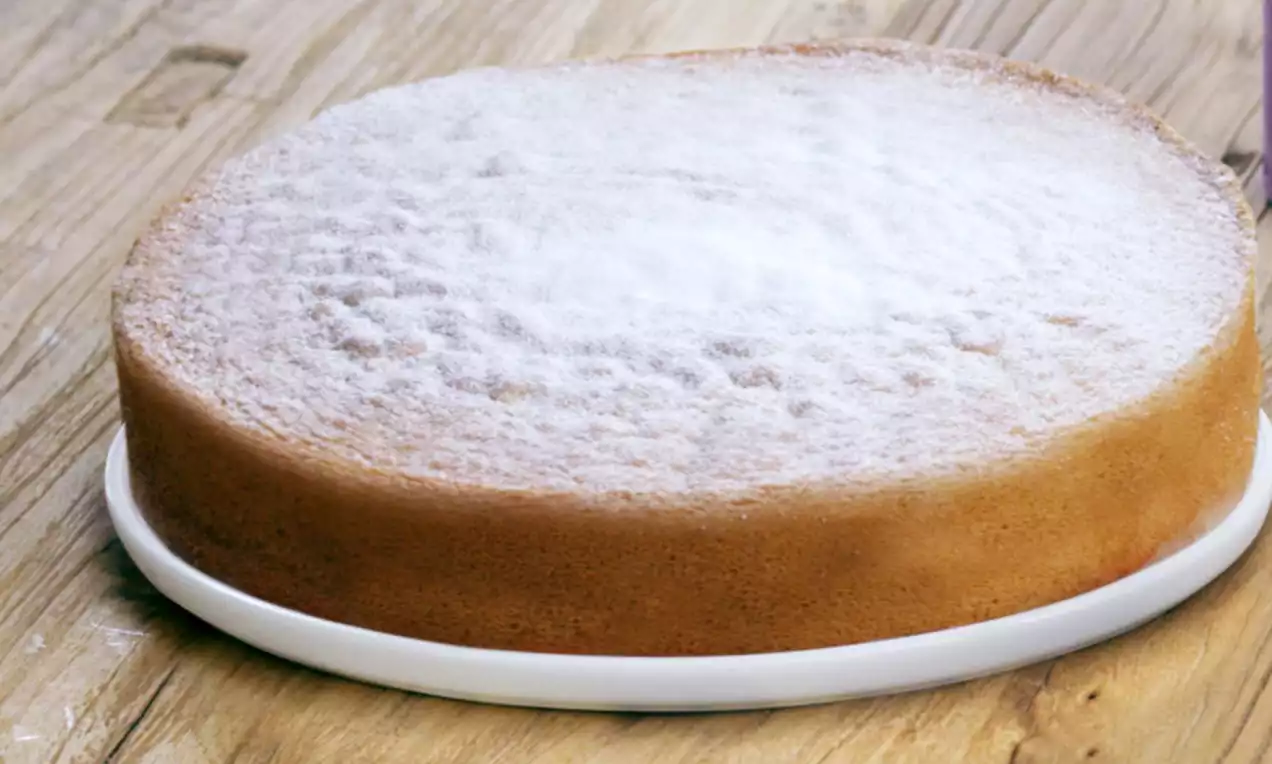 Ronde citroencake met een lichtbruine korst en poederachtige witte laag bovenop, geserveerd op een wit bord op een houten tafel