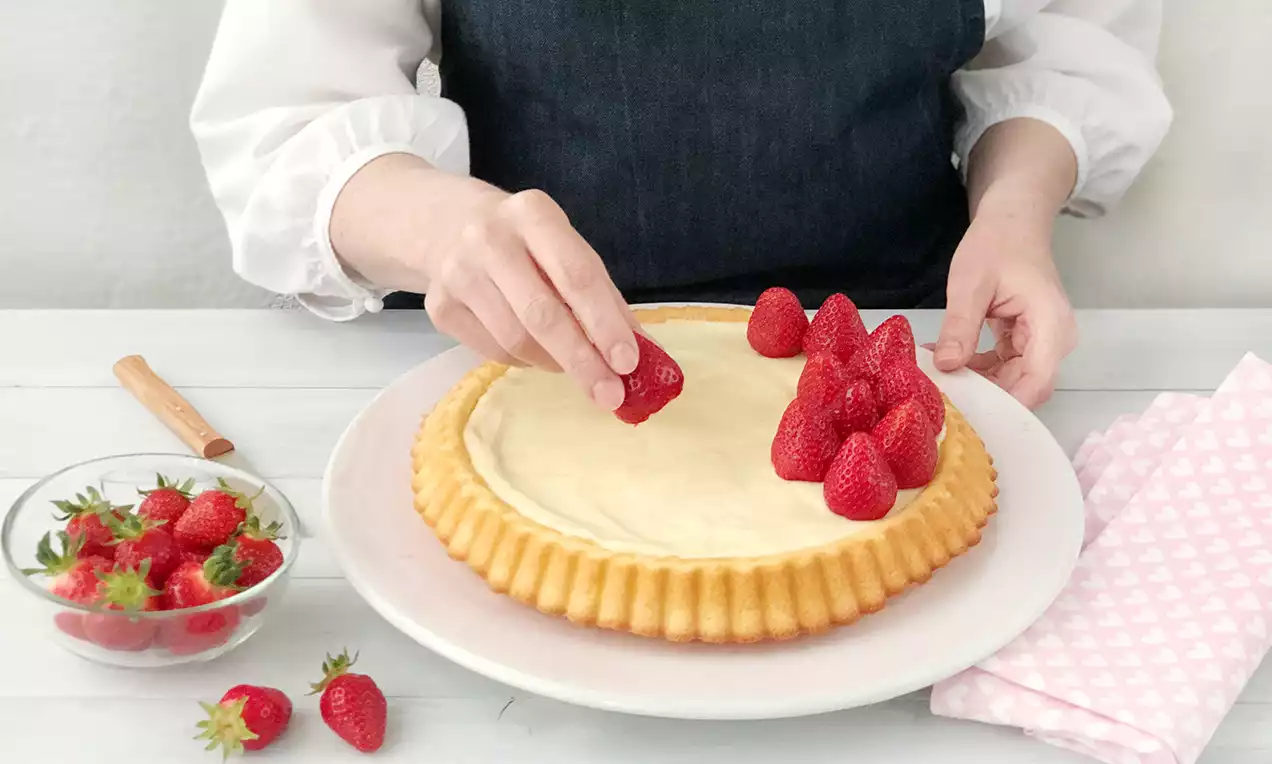 Goldener Tortenboden mit glatter heller Creme, frische rote Erdbeeren werden von Hand darauf platziert, daneben eine Glasschale mit Erdbeeren