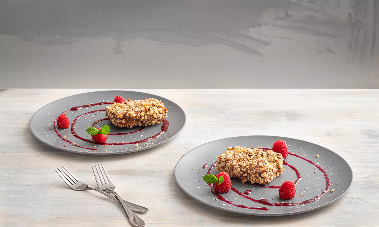 Правоъгълни бисквити с крем, оваляни в златисти ядки, подредени върху сиви чинии с малини, зелени листа и малинов сироп