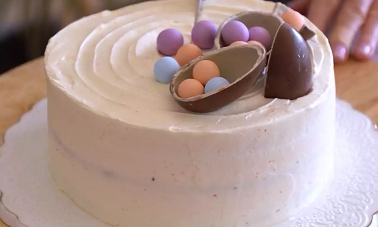 Okrugla torta glatko premazana svijetlom kremom, na vrhu čokoladna jaja s pastelnim kuglicama, na bijelom ukrasnom tanjuru