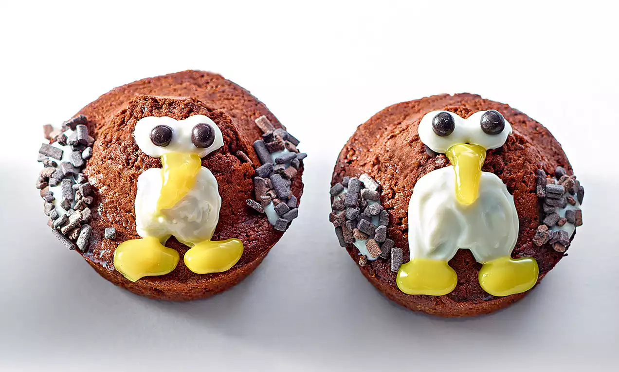 Muffins au chocolat brun décorés en forme de pingouins avec glaçage blanc brillant, bec jaune et éclats chocolatés sur les côtés