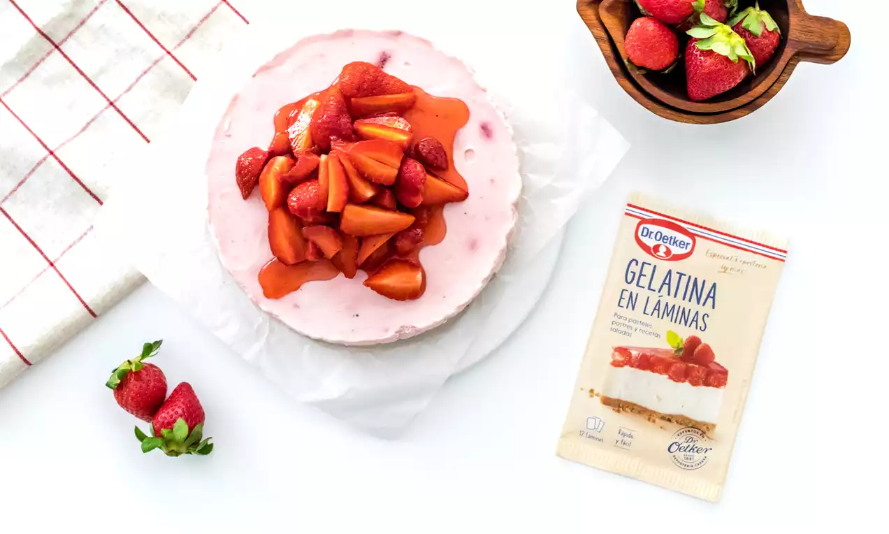 Tarta circular de queso con textura suave y tono rosado, coronada con fresas troceadas y almíbar rojo brillante sobre un plato blanco