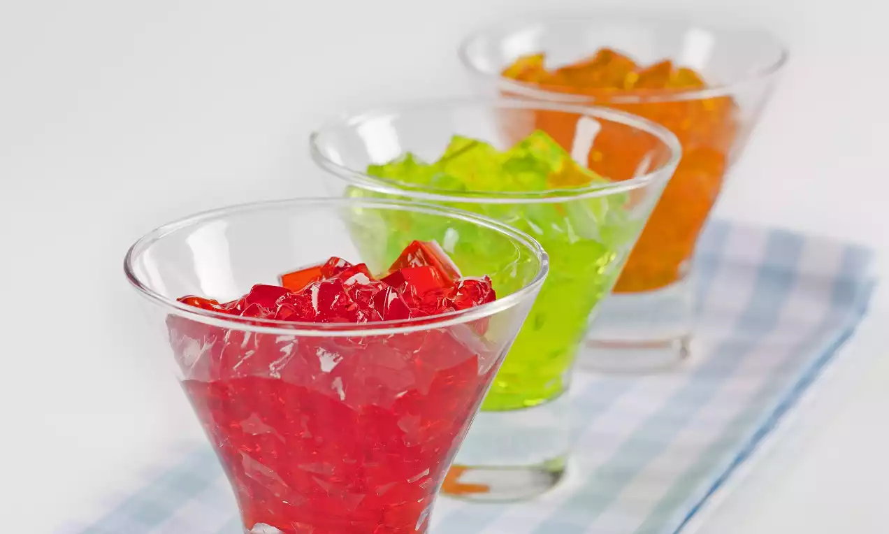 Şeffaf camda sunulan kırmızı, yeşil ve turuncu renkte ışıl ışıl jel küpler, desenli açık mavi örtü üzerinde sıralanmış