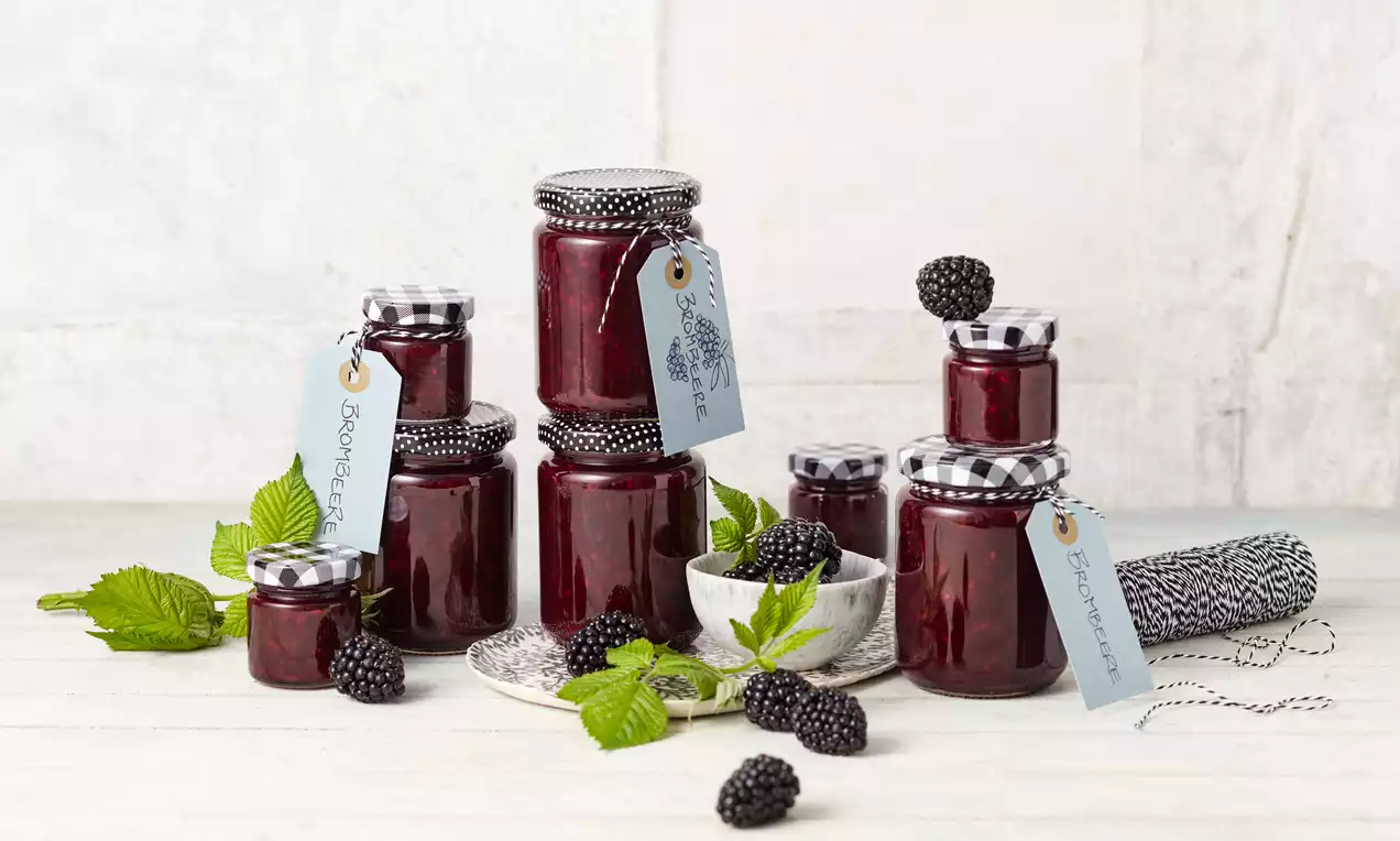 Gläser mit tiefroter Brombeermarmelade, schwarz-weiß karierten Deckeln, blauen Anhängern, frischen Brombeeren und grünen Blättern auf hellem Holz