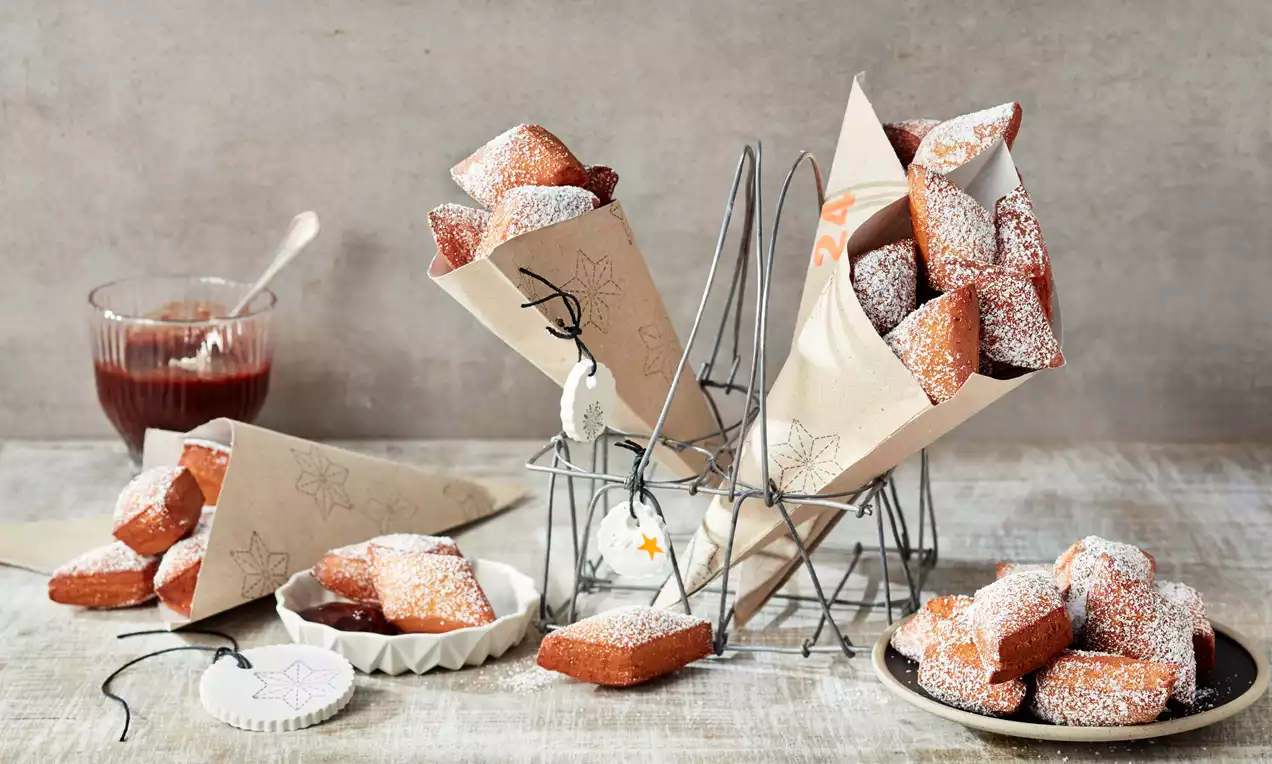 Goldbraune, rechteckige Schmalzkuchen mit puderzuckerbestäubter Oberfläche in Papierhörnchen und auf Tellern, daneben rote Marmelade