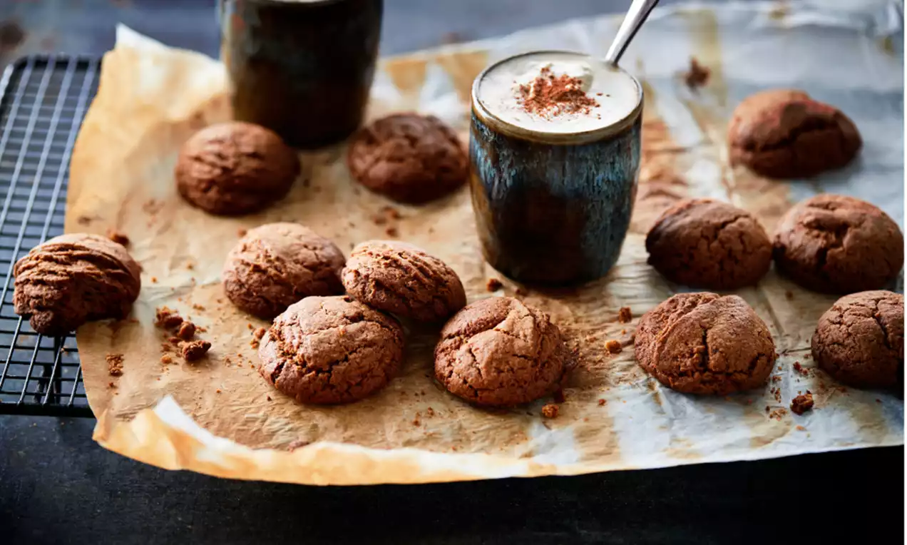 Speculaaskoekjes met ruwe textuur en warme bruine tinten op bakpapier naast rustieke mokken met schuimige koffie en een metalen rooster