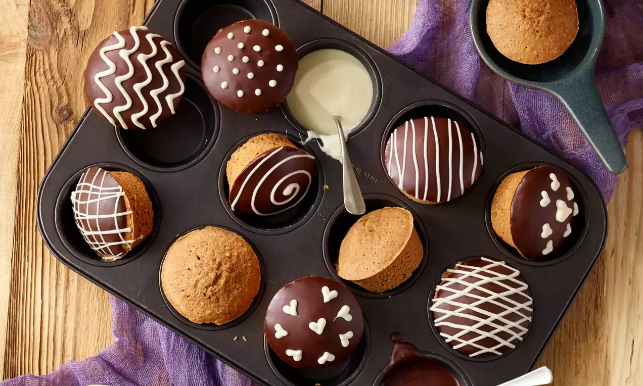 Goldbraune Nuss-Nougat-Muffins, teils mit glänzend dunkler Glasur und filigranen weißen Mustern, arrangiert in einer schwarzen Muffinform