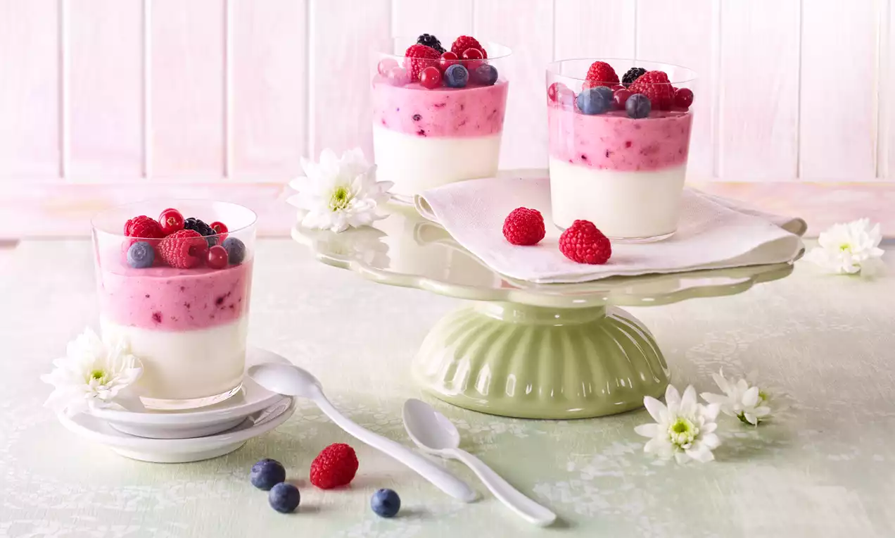 Beeren-Joghurt-Quark-Creme mit rosafarbener Schicht und weißen Unterlagen, dekoriert mit frischen Beeren auf grüner Tortenplatte mit weißen Blumen