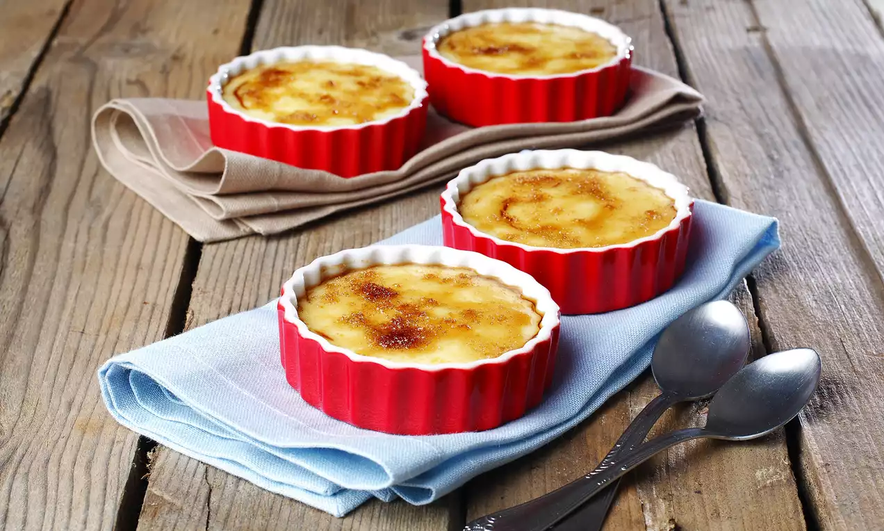 Kırmızı tırtıklı seramik kaplarda sunulan, üstü karamelize edilmiş, kremsi dokulu creme brulee rustik bir ahşap yüzeyde bezlerle süslenmiş