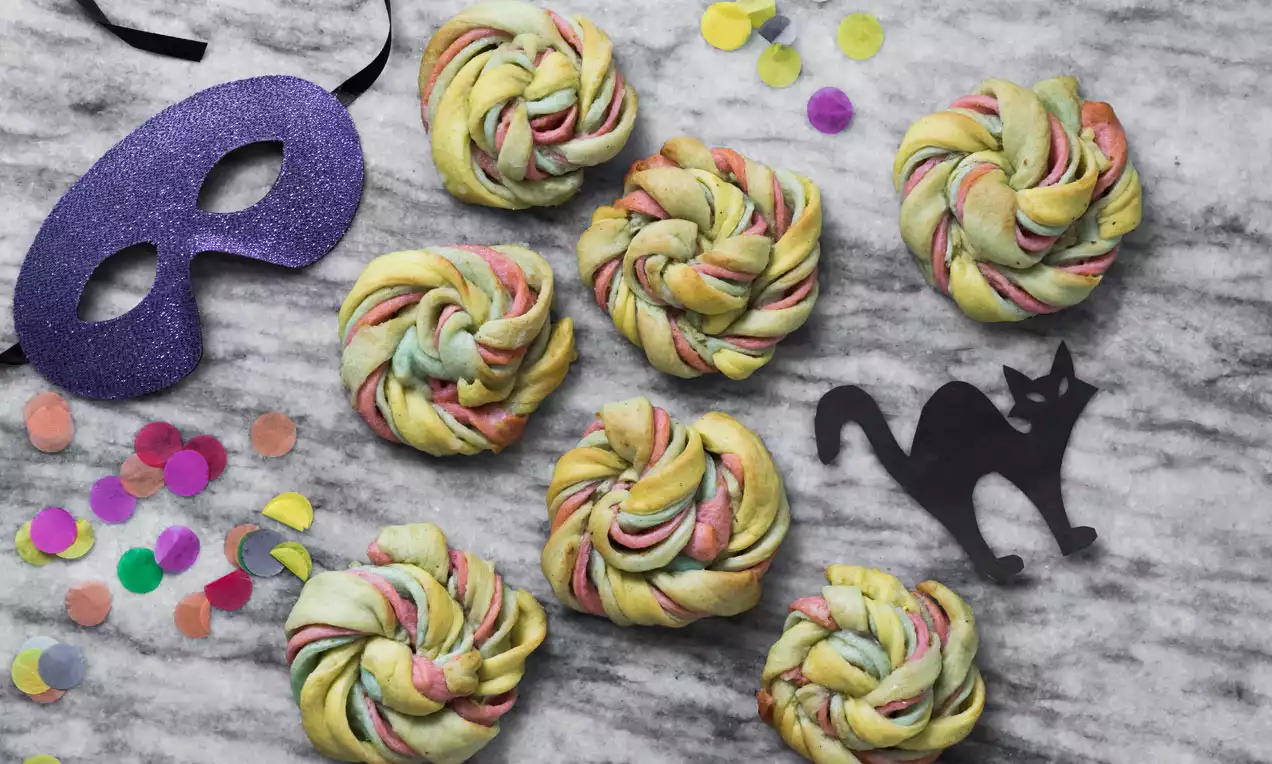 Fargerike fastelavnsboller med flettet spiralform i gul, rosa og grønn deig på en grå marmorbunn pyntet med konfetti, maske og svart katt