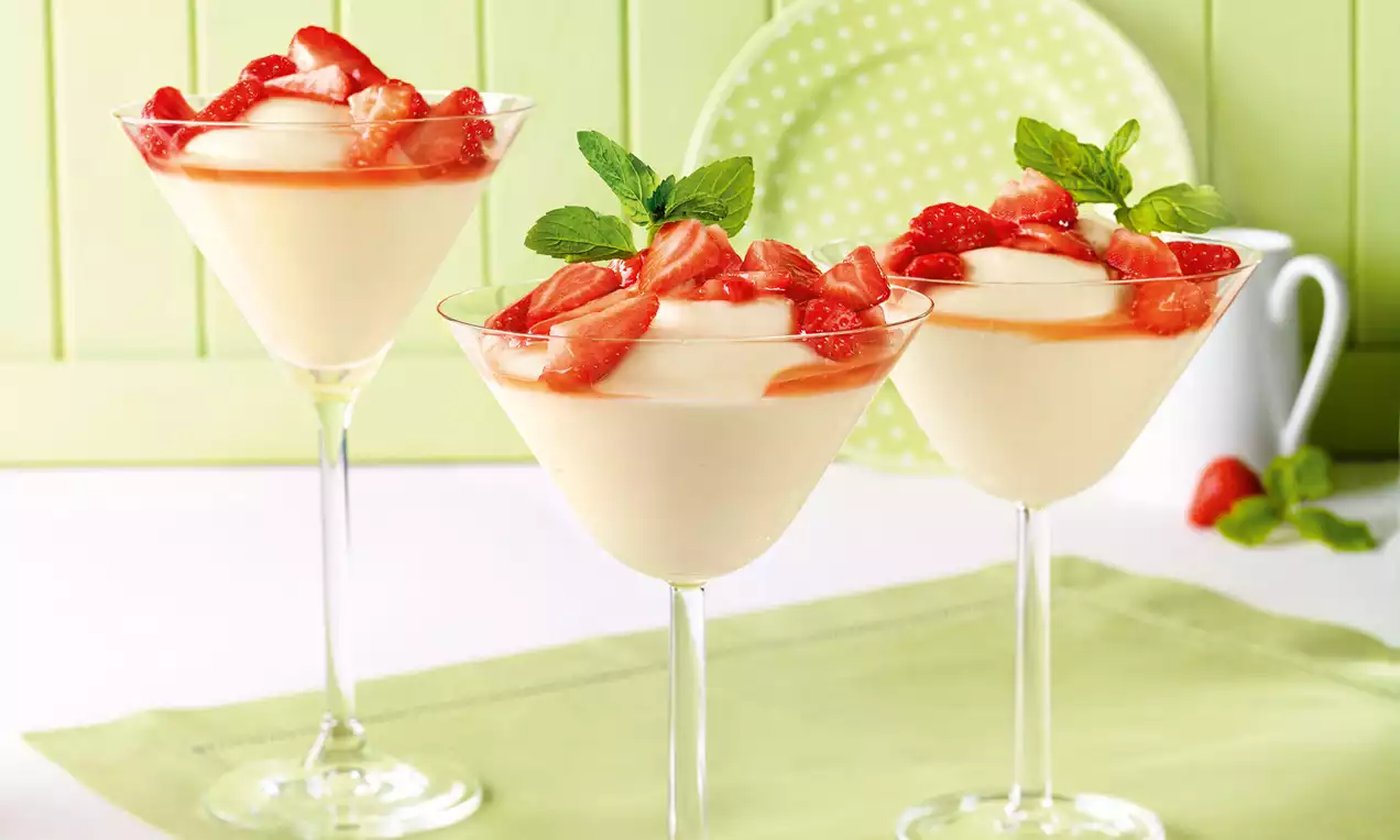 Kremast bezgov puding bledo rumene barve v steklenih kozarcih, okrašen s svežimi rdečimi jagodami in listi mete na svetlo zelenem prtu