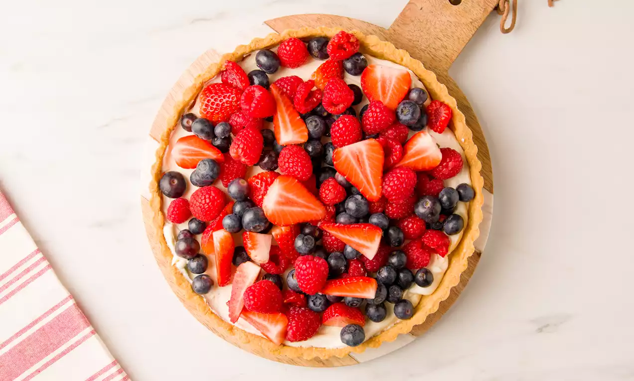 Tartaleta de masa dorada rellena de crema blanca de mascarpone, cubierta con fresas, frambuesas y arándanos sobre tabla de madera