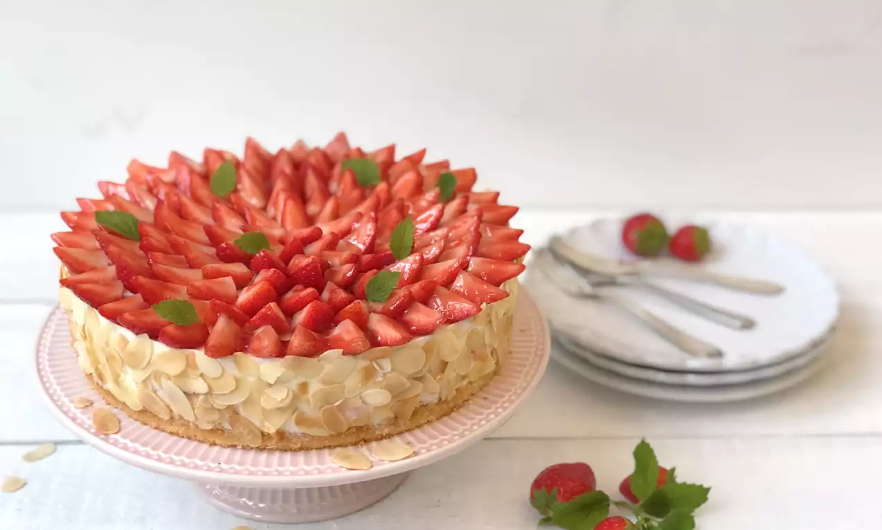 Erdbeer Quark Torte mit Mandelsplittern am Rand, dicht belegten roten Erdbeerscheiben und grünen Minzblättern auf rosa Tortenplatte
