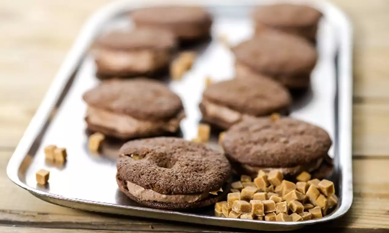 Suklaanruskeat, pyöreät whoopiet pehmeällä täytteellä tarjottimella, vierellä pieniä vaaleanruskeita fudge-kuutioita