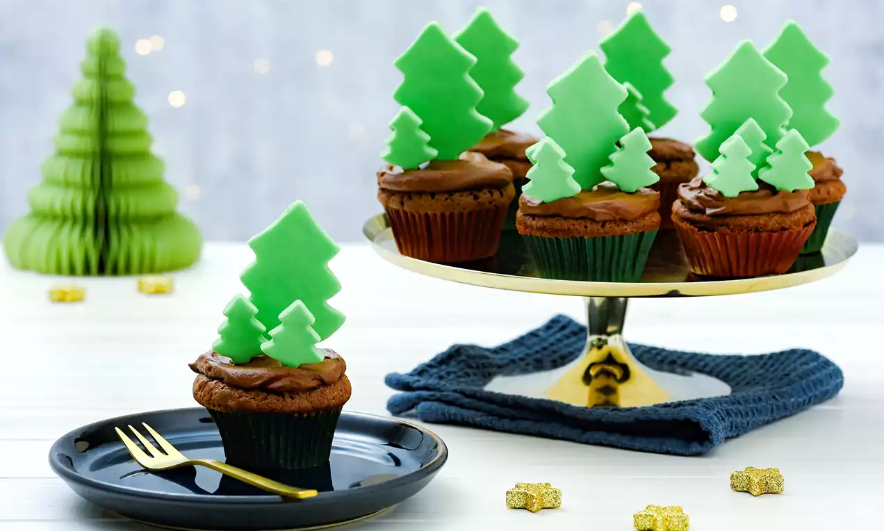 Chocolate cupcakes topped with glossy frosting and green tree-shaped decorations on a gold cake stand with festive accents