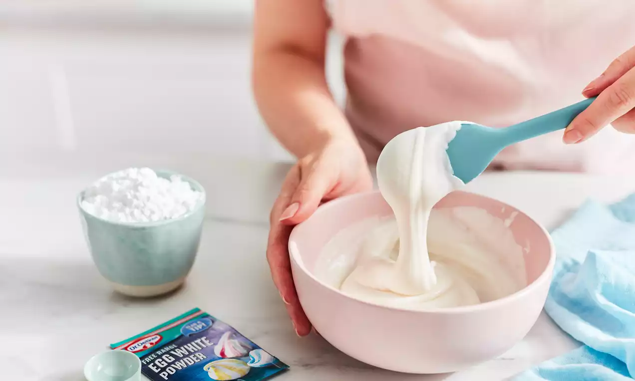 Glossy white royal icing with a smooth, creamy texture being stirred with a blue spatula in a pale pink bowl on a marble countertop