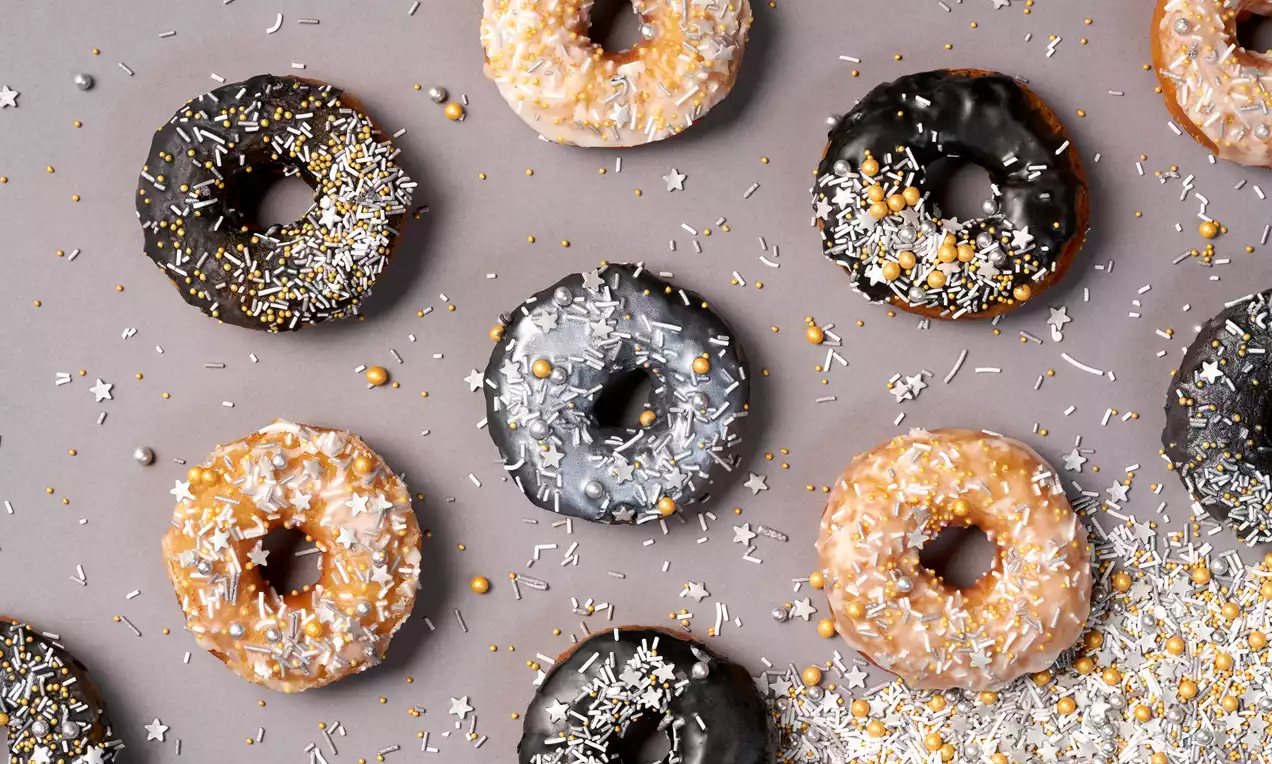 Donkere ronde donut met glanzende chocoladelaag en kleurrijke sprinkles op een grijze ondergrond met verspreide stervormige decoraties