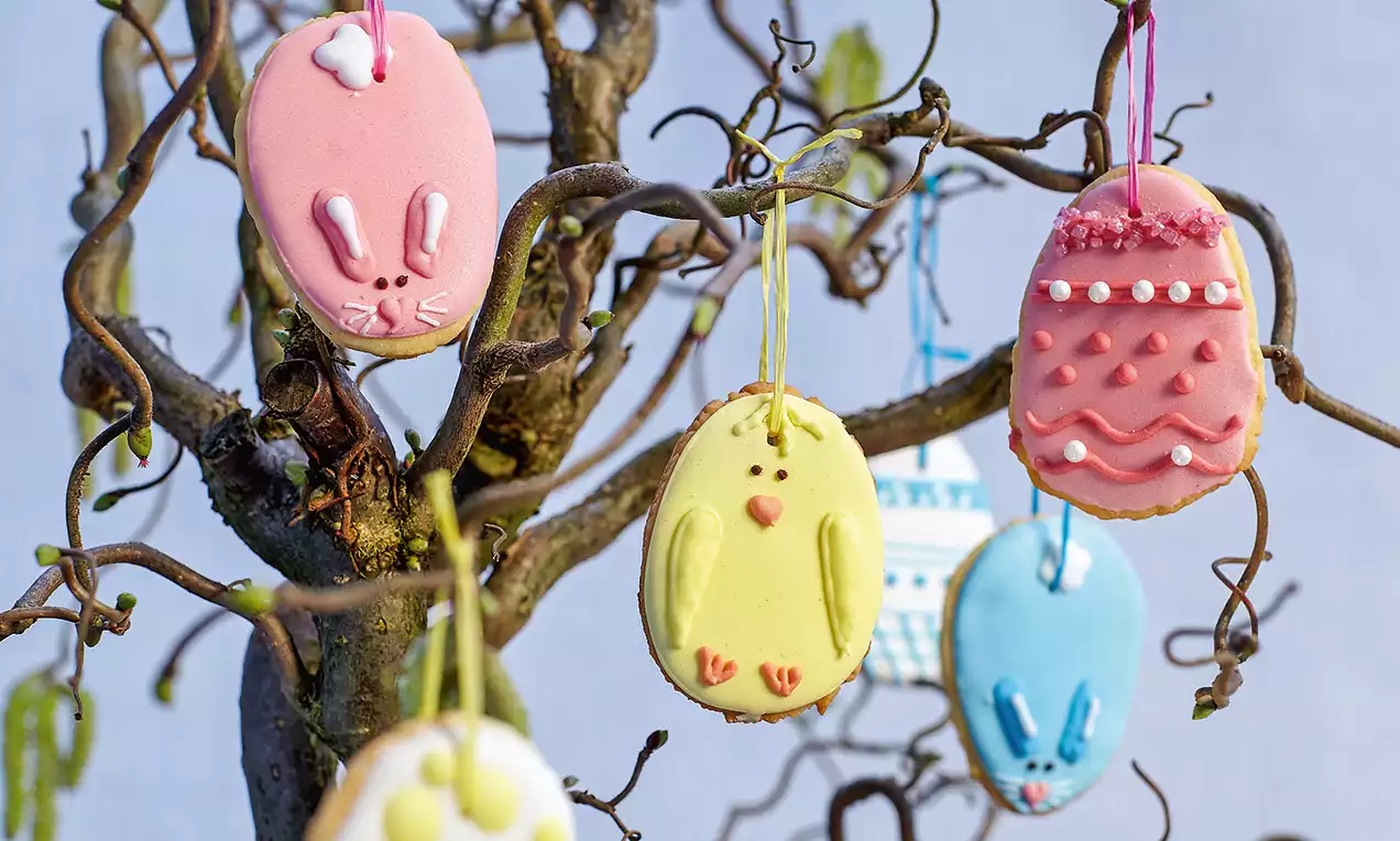 Biscuits en forme d'œufs colorés suspendus à un arbre tortueux, décorés de motifs lapins, poussins et perles en glaçage