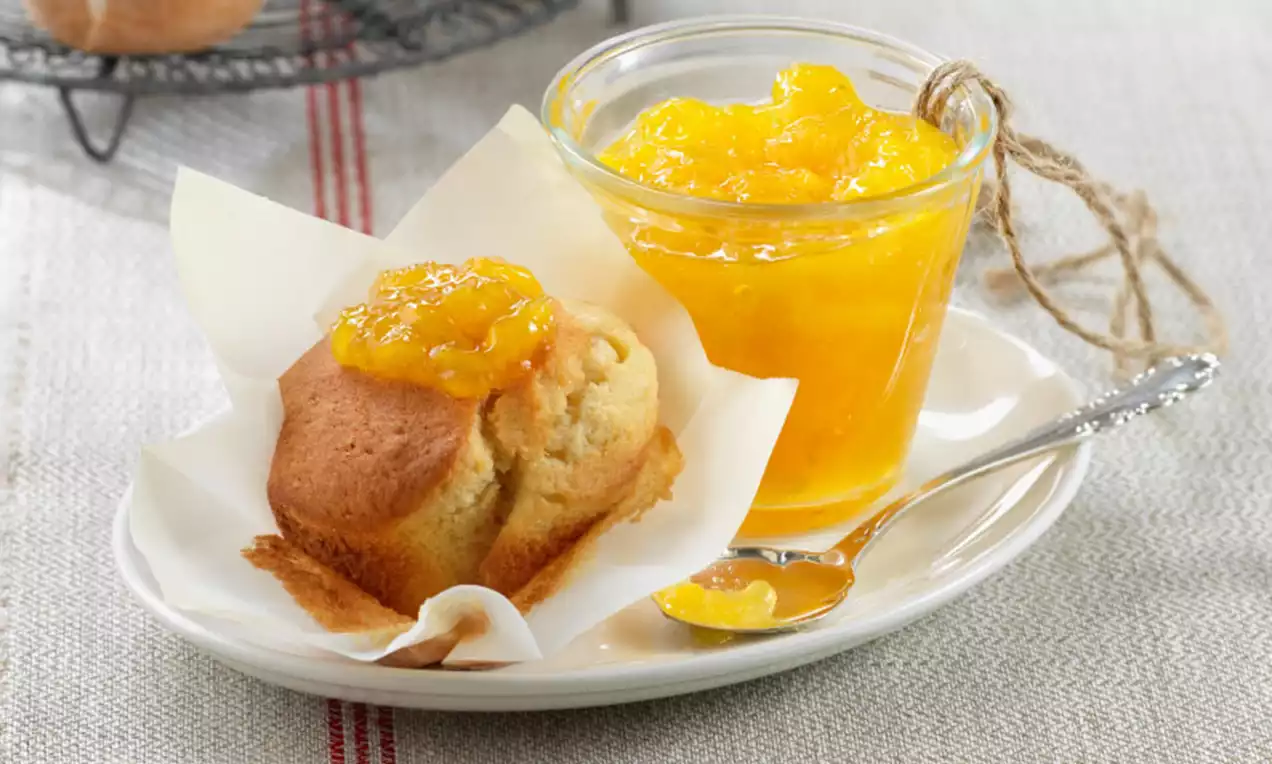 Mermelada brillante de mango amarillo anaranjado en un vaso transparente junto a un bizcocho dorado servido sobre un plato blanco con cucharilla