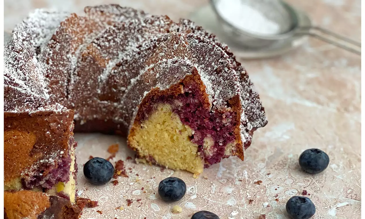 Marmorierter Gugelhupf mit goldbraunem Rand, Blaubeer-Swirl und Puderzucker, daneben frische Blaubeeren und ein Metallsieb