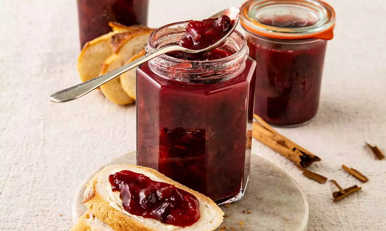 Confiture Zwetschgen-Zimt rouge profonde dans un pot hexagonal, texture lisse et brillante avec des tranches de pain doré et bâtons de cannelle