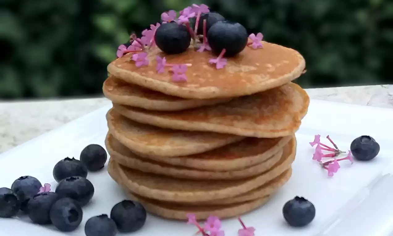 Na bílém talíři je věž ze zlatavých lívanců s měkkou texturou, zdobená temně modrými borůvkami a drobnými růžovými kvítky