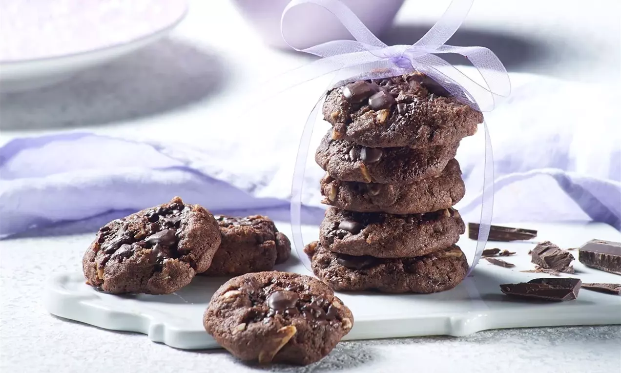 Кафяви веган бисквити с неравна повърхност, едни подредени на купчинка с лилава панделка, други разпръснати върху бяла дъска