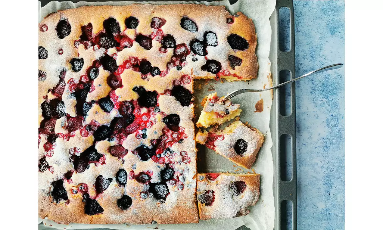 Rektangulær gyllenbrun bærkake på bakepapir, toppet med mørke bær og melisdryss, flere oppskårne stykker med serveringsskje