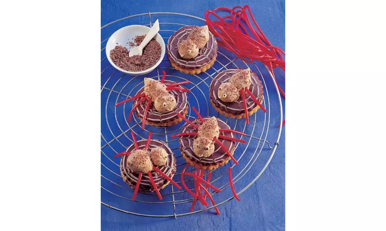 Petits gâteaux ronds chocolatés sur une grille argentée, garnis de corps en biscuits et pattes en fil rouge sur fond bleu