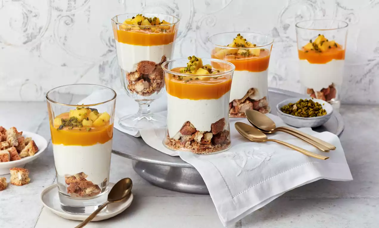 Gläser mit weißer Creme auf Keksstücken, darüber leuchtend gelber Mangospiegel mit Mangowürfeln und Pistazien, arrangiert auf silbernem Teller