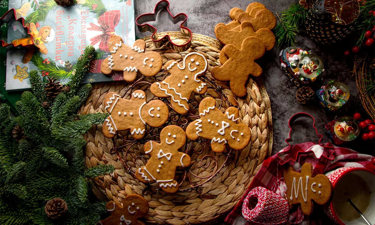 Подредени върху плетена подложка джинджифилови бисквитки в топъл кафяв цвят с бели захарни орнаменти до формички и зимна украса