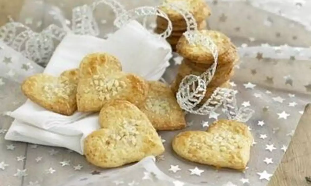 Hjerteformede, gyllenbrune småkaker med hakkede nøtter på en serviett, omgitt av stjernemønstret stoff og dekorative sølvbånd