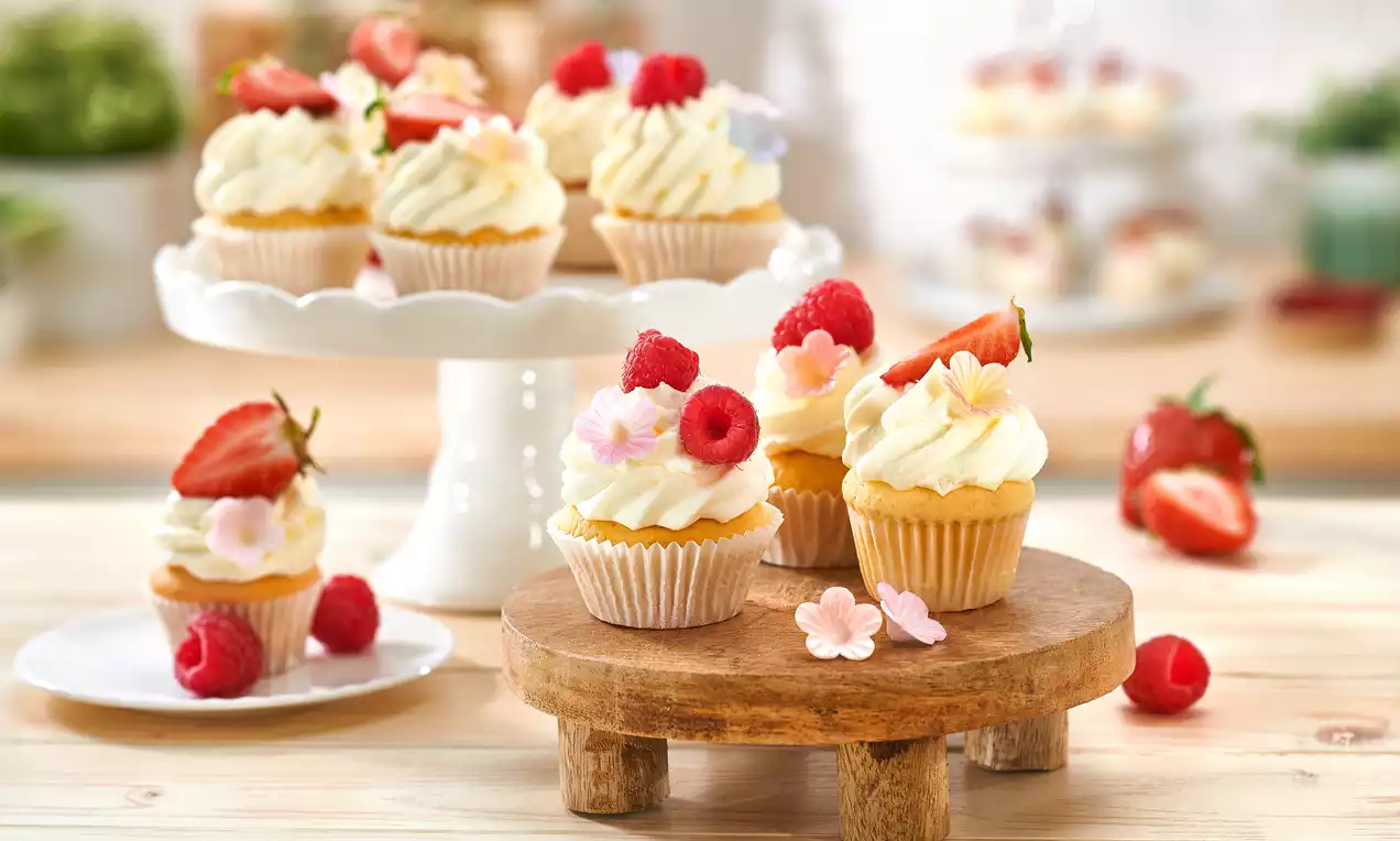 Mini-Cupcakes mit goldgelbem Boden und weißer Cremetopping, garniert mit roten Beeren und Blumen auf Holzplatte und Tortenständer drapiert