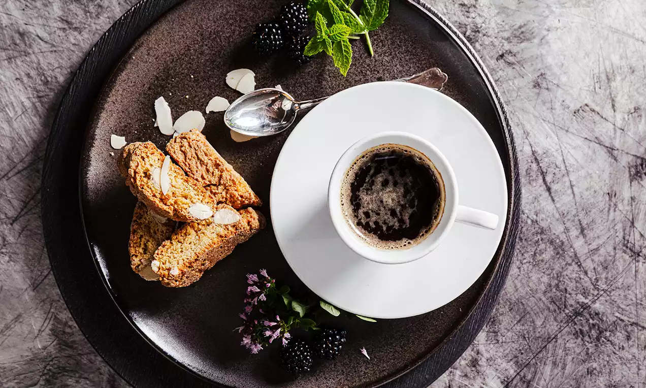 Brun tallrik med gyllenbruna skivade biscotti bredvid en vit kaffekopp, dekorerad med björnbär, mandelspån, gröna blad och blommor