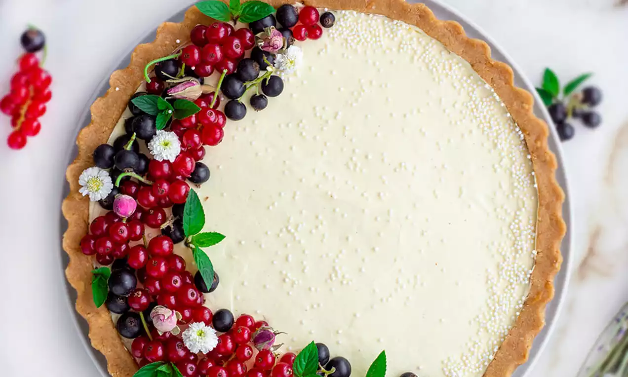 Nepečeni tart s kremastim svijetložutim punjenjem u glatkoj hrskavoj korici, ukrašen crvenim i crnim bobicama te listićima mente