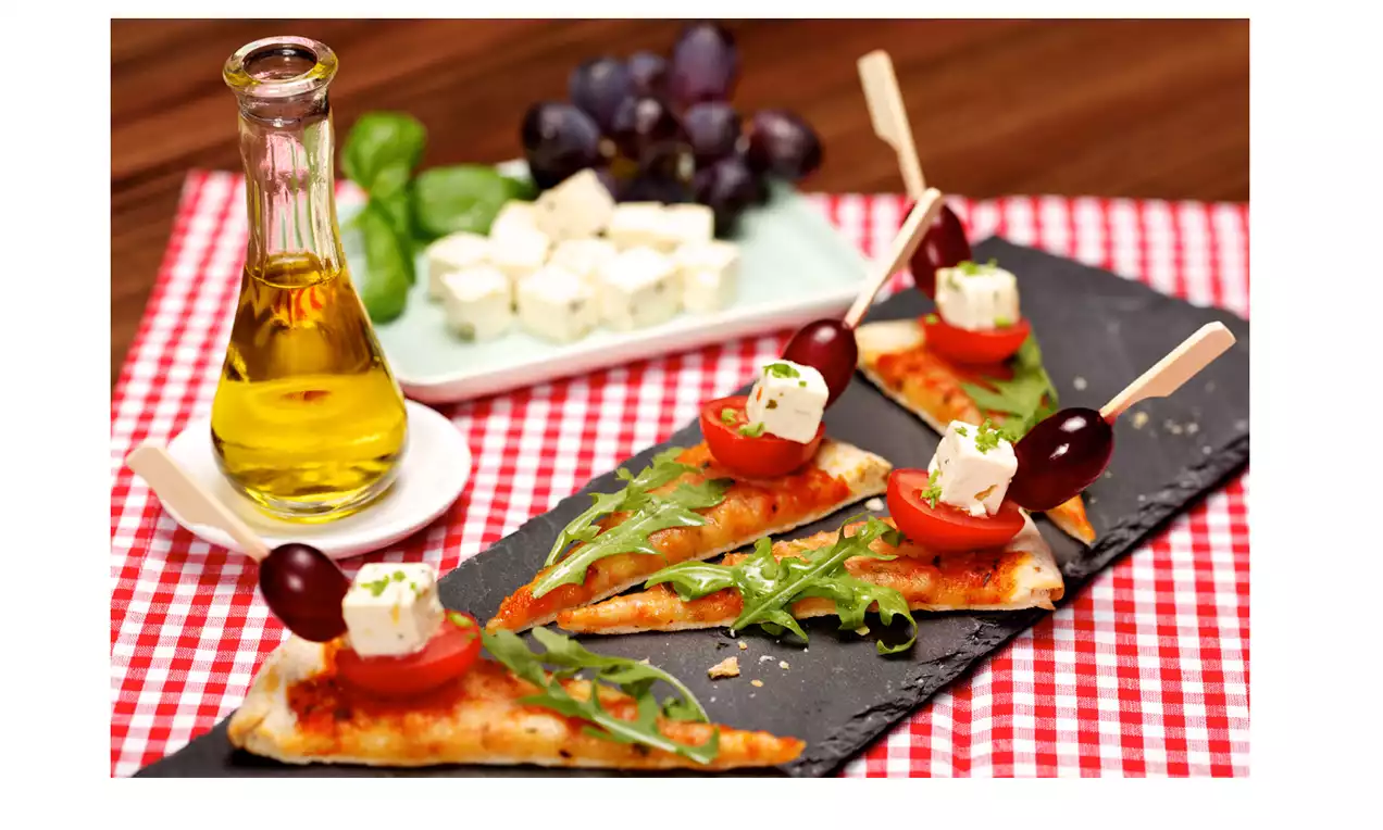 Triangles de pizza dorés garnis de roquette, tomates cerises, cubes de feta et olives noires, présentés sur une ardoise noire