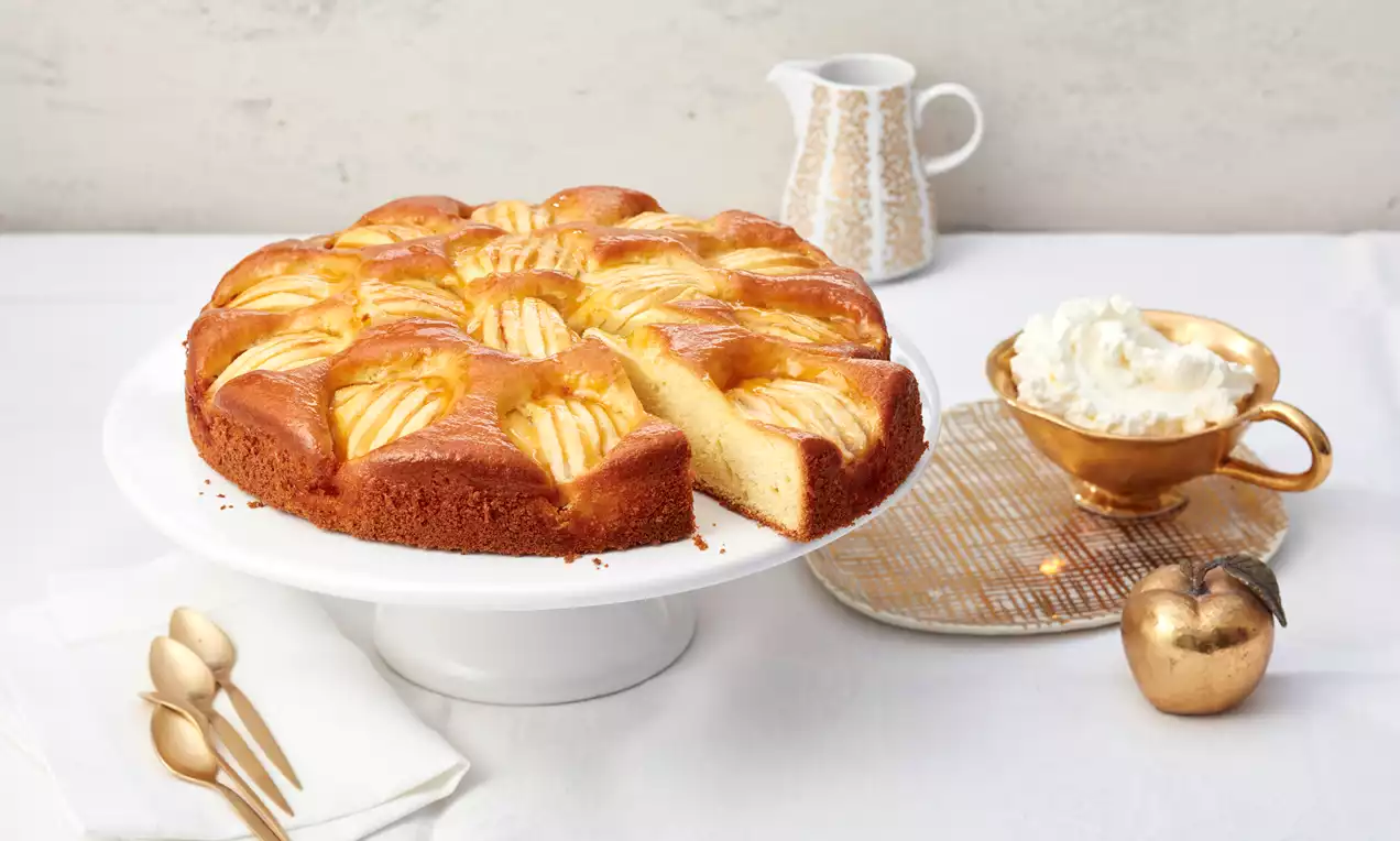 Apfelkuchen mit goldbrauner Kruste und üppig angeordneten glänzenden Apfelscheiben, angeschnittene Portion zeigt saftigen hellen Rührteig