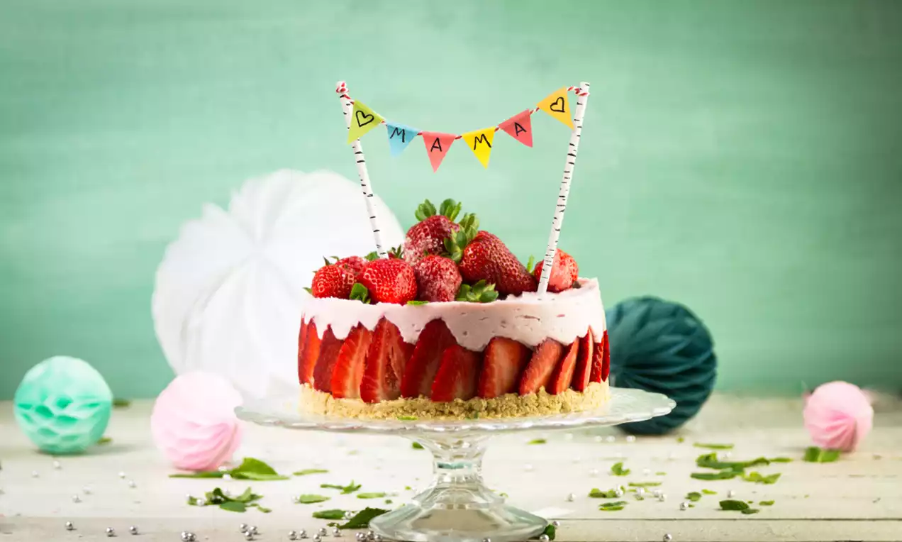 Fruityoghurttaart met roze crème, aardbeien bovenop en langs de rand, op een glazen schaal met speelse vlaggetjes en groene decoratie