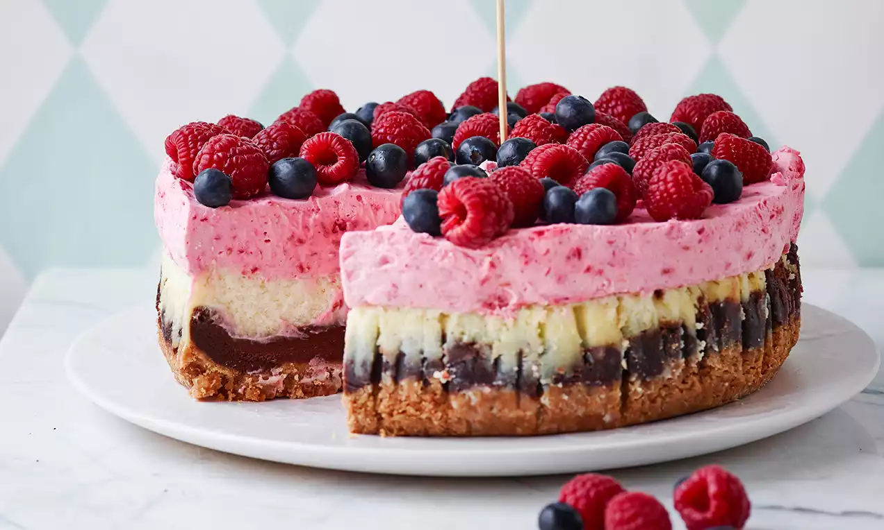 Kage med lyserød bærtopping, lag af creme og chokolade ovenpå en gylden kiksebund, dekoreret med friske bær på hvid tallerken